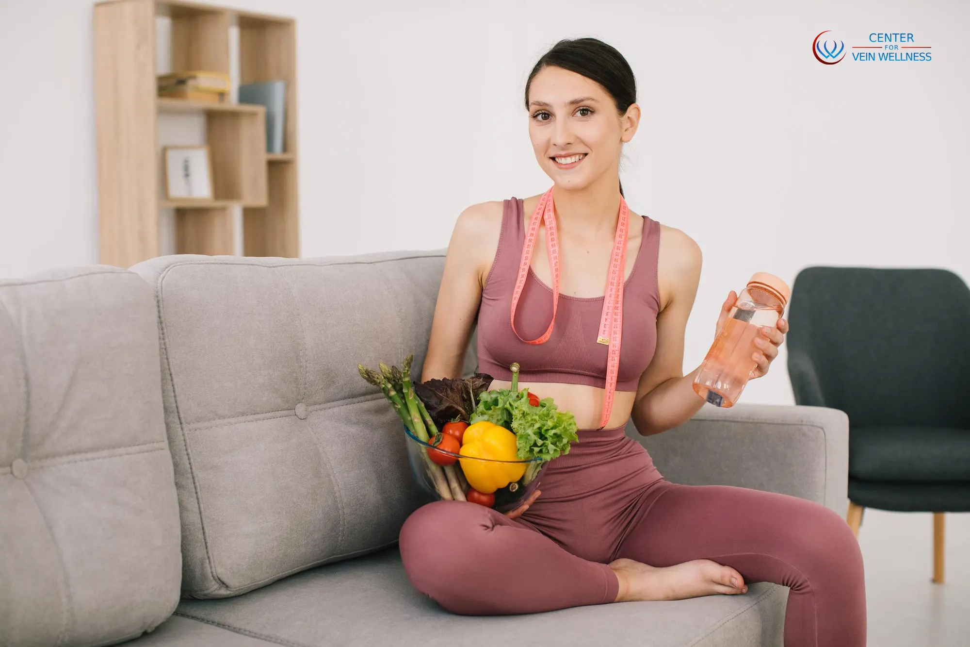 Young woman in athletic wear sitting on a sofa, smiling, and holding a bowl of fresh vegetables and a bottle of water. She has a pink measuring tape around her neck, emphasizing a healthy lifestyle. A logo in the corner reads 'Center for Vein Wellness